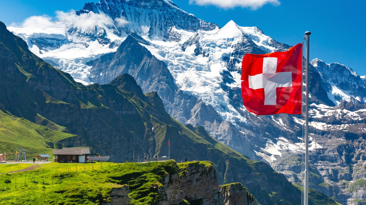 A stunning Swiss Alpine landscape featuring a vibrant red Swiss flag, snow-capped mountains, and lush green pastures, symbolizing the purity and heritage of Swiss-grown herbs